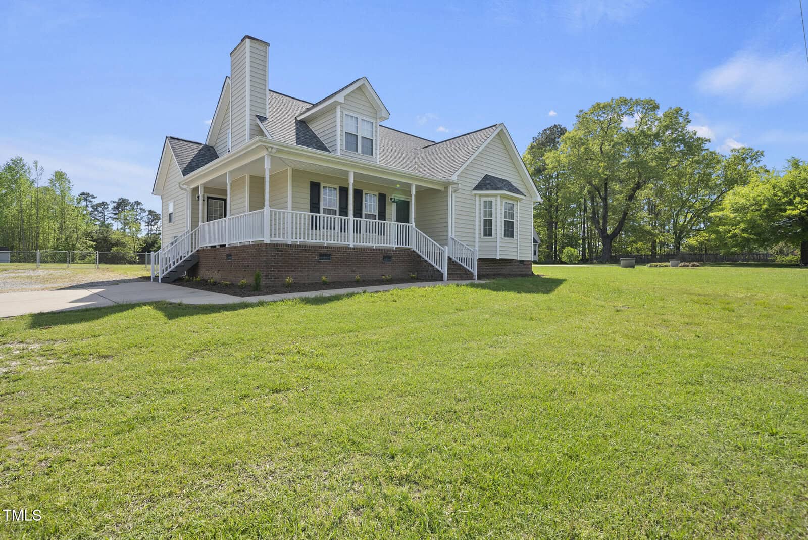 A beige two-story house with a covered front porch, brick foundation, and a driveway, situated on a large grassy lawn with trees in the background on a sunny day.