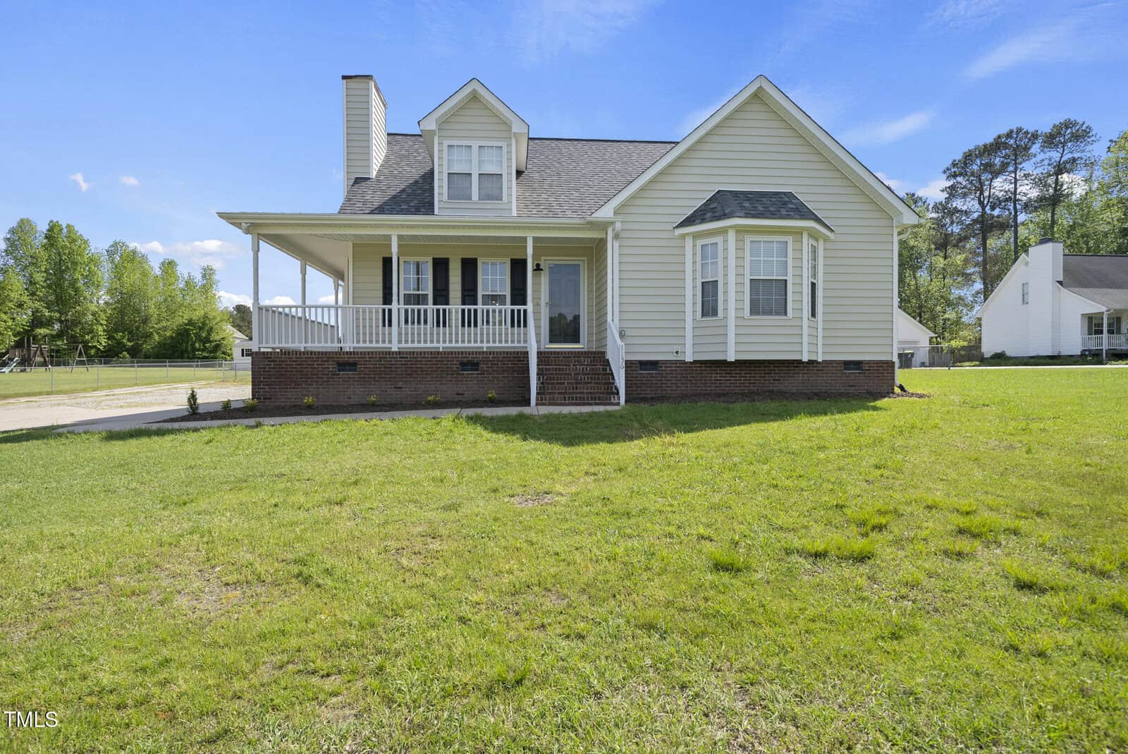 A light yellow single-family house with a brick foundation, front porch, and dormer windows, sits on a grassy lawn under a blue sky with trees and another house in the background.