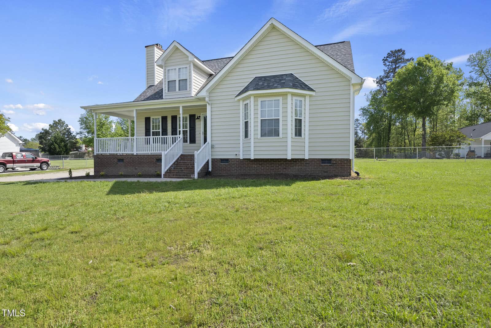 A beige two-story house with white trim, a covered front porch, brick foundation, and a bay window, situated on a large grassy lawn with trees and a fenced yard in the background.