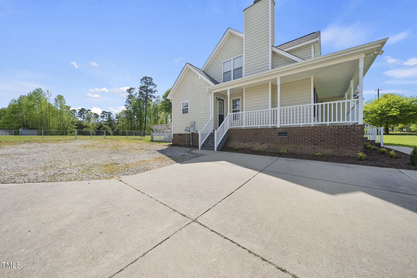 A two-story beige house with white trim, a large covered front porch, brick foundation, and concrete driveway. The yard has a gravel section and is bordered by grass and trees under a blue sky.