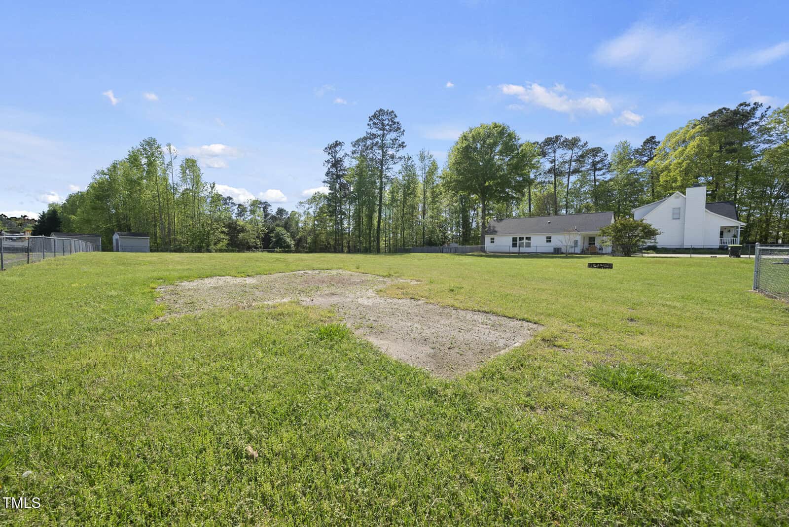 A grassy backyard with a rectangular patch of bare dirt in the foreground, surrounded by trees and a white single-story house with a fenced yard in the background under a blue sky with scattered clouds.