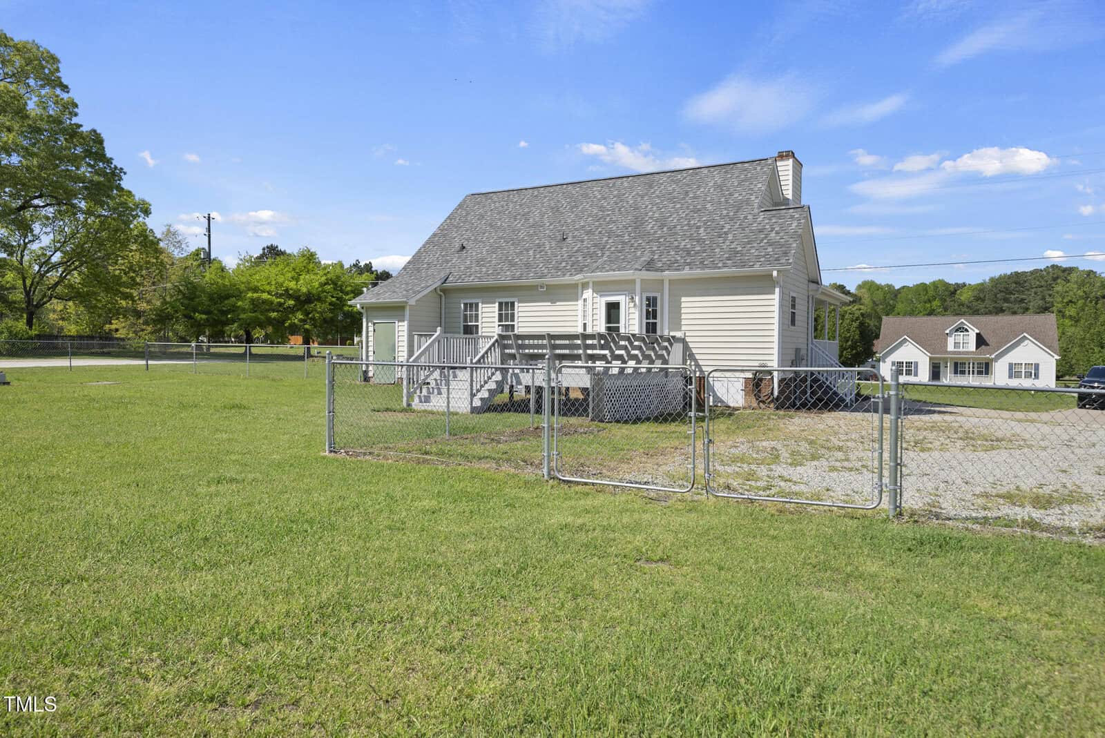 A small beige house with a gray roof and back deck sits in a grassy yard enclosed by a chain-link fence. Another house and trees are visible in the background under a blue sky with clouds.