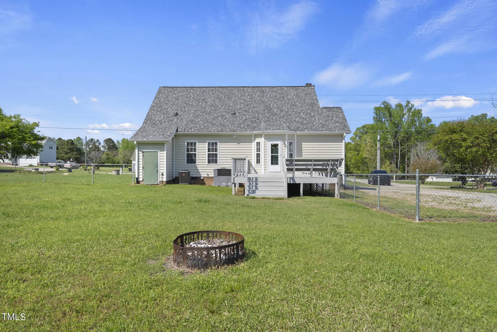 A house with light siding, a dark shingled roof, small porch steps, and a fenced backyard with a round metal fire pit on grass. There are trees, a driveway, and blue sky with clouds in the background.