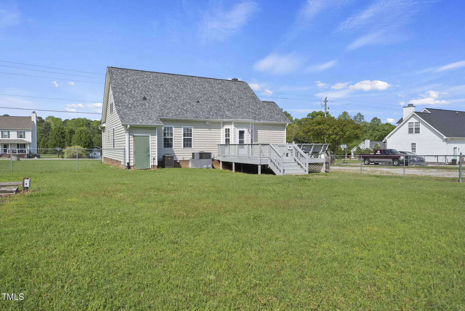 Single-story house with gray siding, a shingled roof, raised wooden deck, and fenced backyard. The yard is grassy and spacious, with neighboring houses and vehicles visible in the background under a blue sky.
