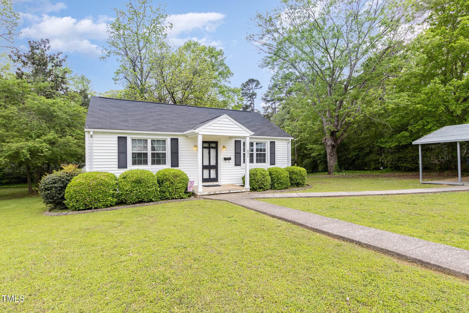 A small white house with black shutters and a black door sits on a green lawn, surrounded by bushes and trees. A concrete walkway leads to the front entrance, and a driveway with a carport is on the right.
