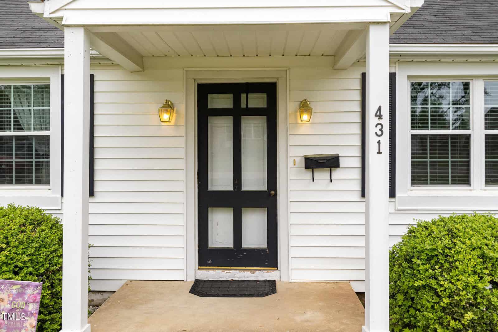 A white house with a covered front porch, black-framed door with glass panels, two lit wall lanterns, black mailbox, house number 431, windows with white trim, and green bushes on either side of the entrance.