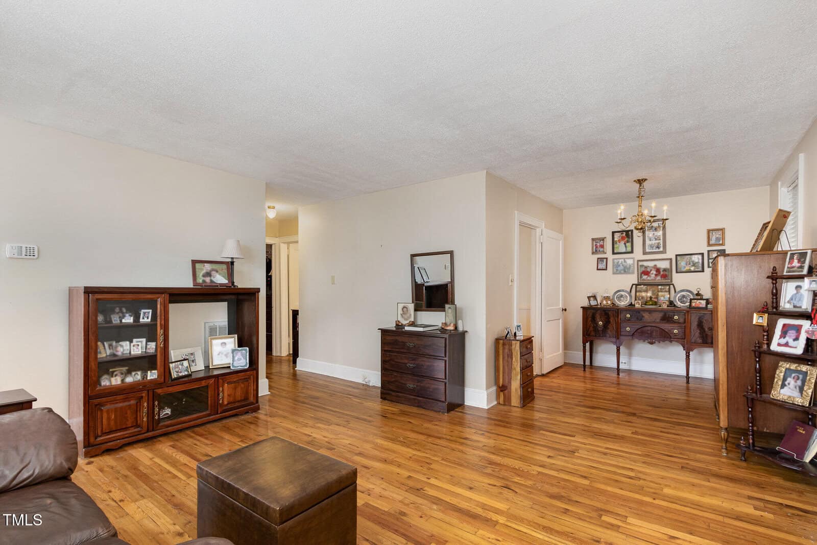 A living room with hardwood floors, cream-colored walls, and brown wooden furniture displaying framed photographs. A chandelier hangs over a sideboard, and there are several doors leading to other rooms.