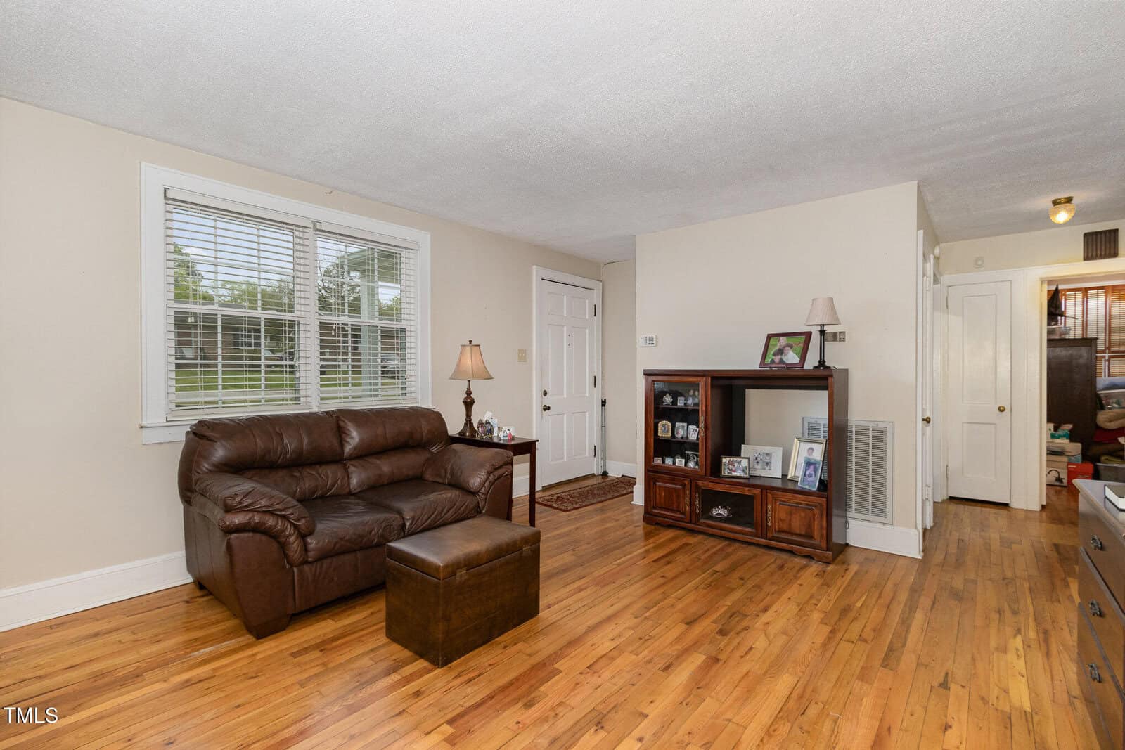 A living room with light-colored walls, hardwood floors, a brown leather loveseat and ottoman, a wooden TV stand with shelves, framed photos, two table lamps, a window, and a door leading outside.