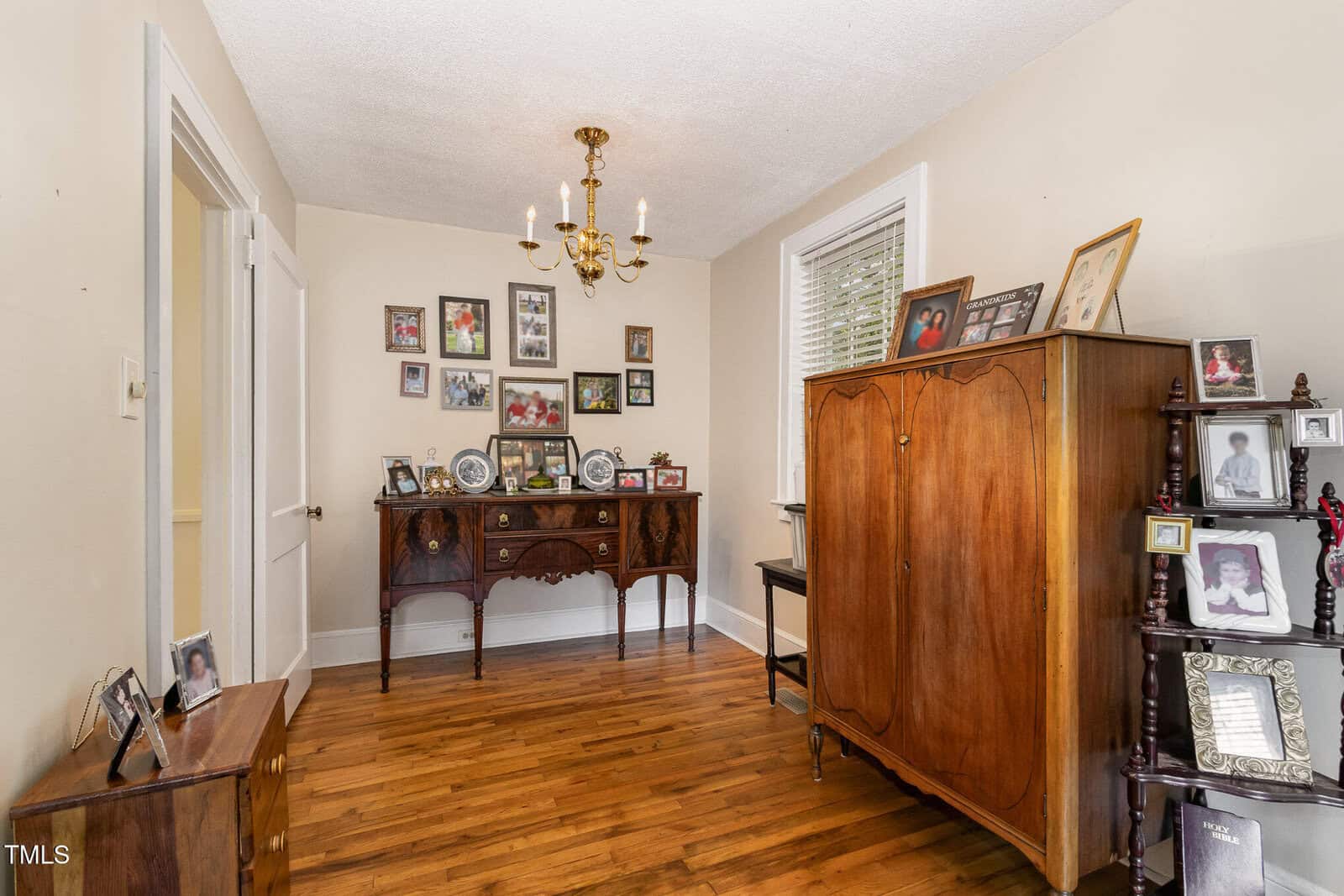 A room with hardwood floors, cream-colored walls, a chandelier, a wooden cabinet, a set of shelves with framed photos, and a sideboard with more framed photographs and decorations.