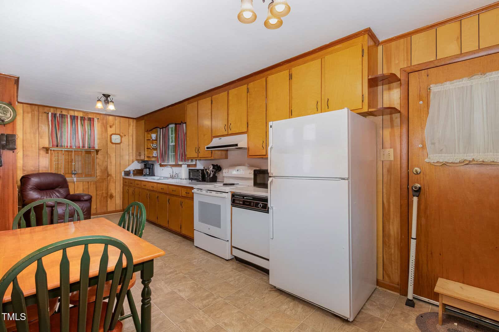 A kitchen with wood-paneled walls, light brown cabinets, a white stove, white refrigerator, microwave, and a wooden dining table with green chairs. A brown door and a small lounging area are also visible.