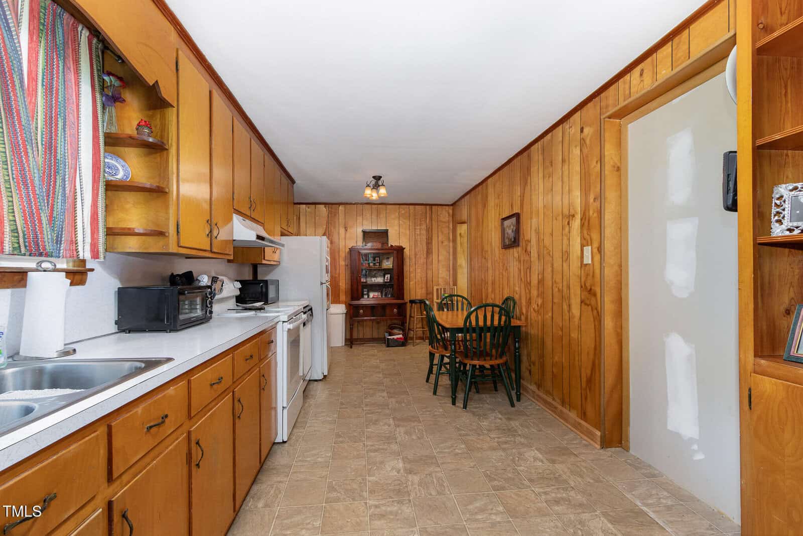 A kitchen with wood-paneled walls and cabinets, white appliances, a double sink, and a small dining area with a round table and four chairs. Shelves and a hutch hold dishes and other items.