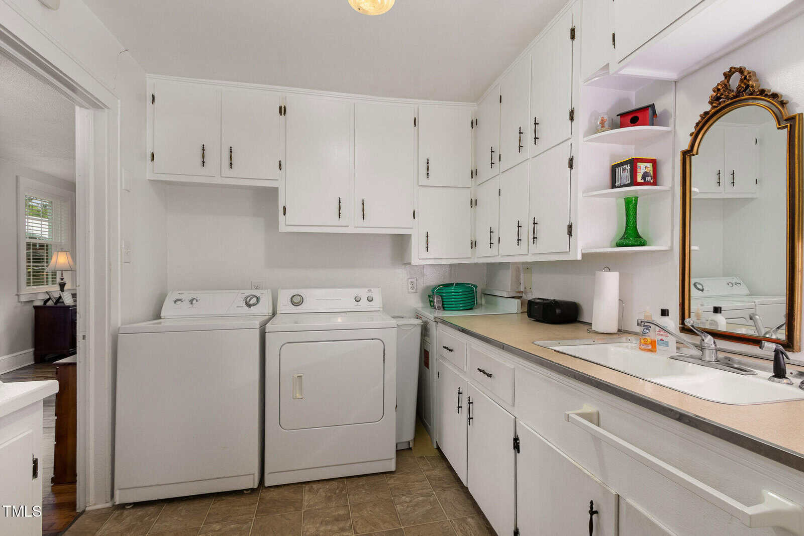 Laundry room with a washing machine and dryer beside a sink, white cabinets above and below, tan countertop, mirror with gold frame, paper towel roll, and various small items on the counter.