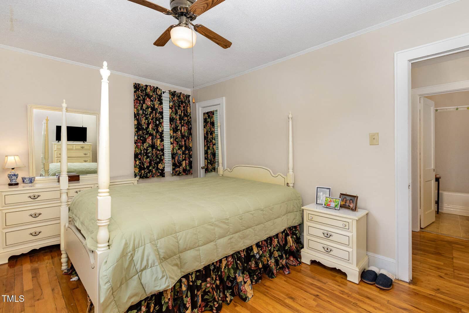 A bedroom with a four-poster bed, green bedspread, two floral curtains, two white nightstands with lamps and framed photos, a ceiling fan, and hardwood floors. A door to a hallway is open.