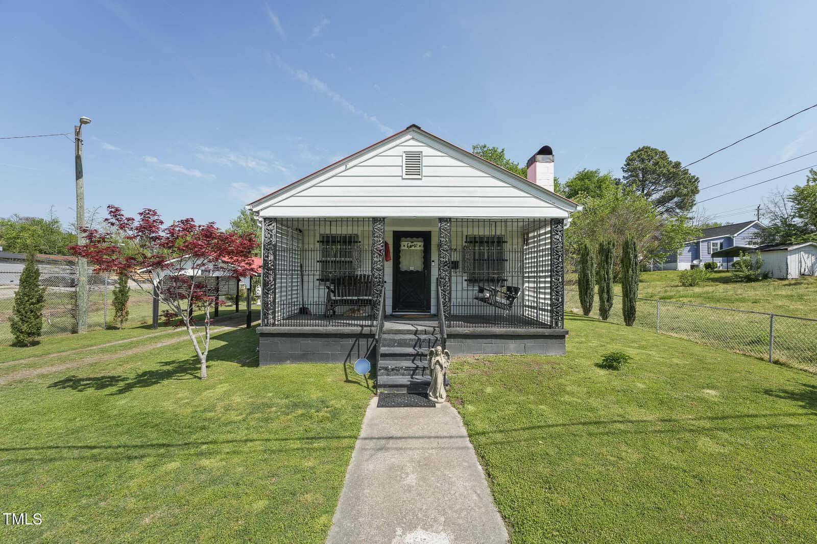 A single-story white house at 729 S Carolina Avenue, Henderson, features a covered front porch with black railing and steps leading to a paved walkway. The grassy yard has small trees, bushes, and a statue near the porch steps.