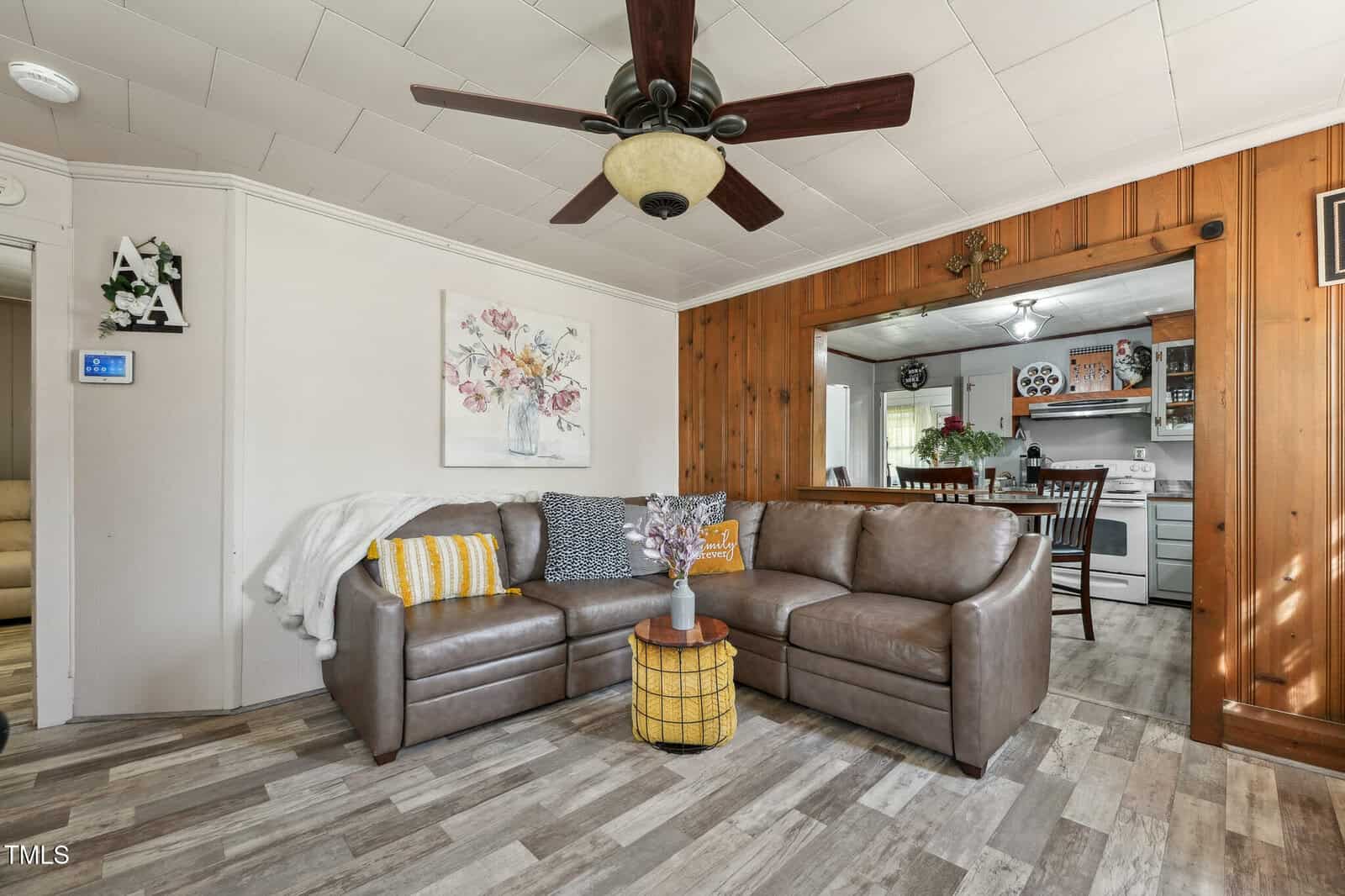 A living room with a brown sectional sofa, two patterned pillows, a throw blanket, a round wooden coffee table, and a ceiling fan. The room has wood-paneled walls and an open view into the kitchen.