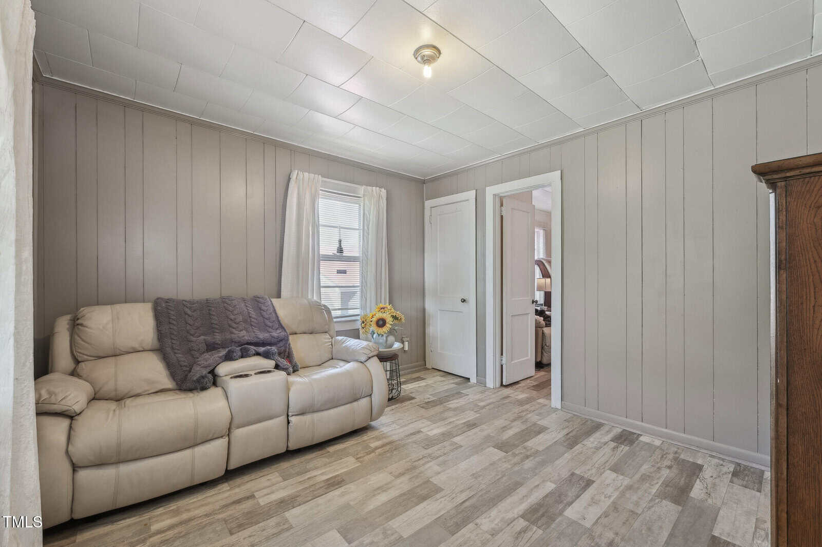 A small living room with light wood flooring, gray paneled walls, a beige recliner sofa with a gray blanket, a side table with sunflowers, and a window with white curtains. Two doors are visible in the room.
