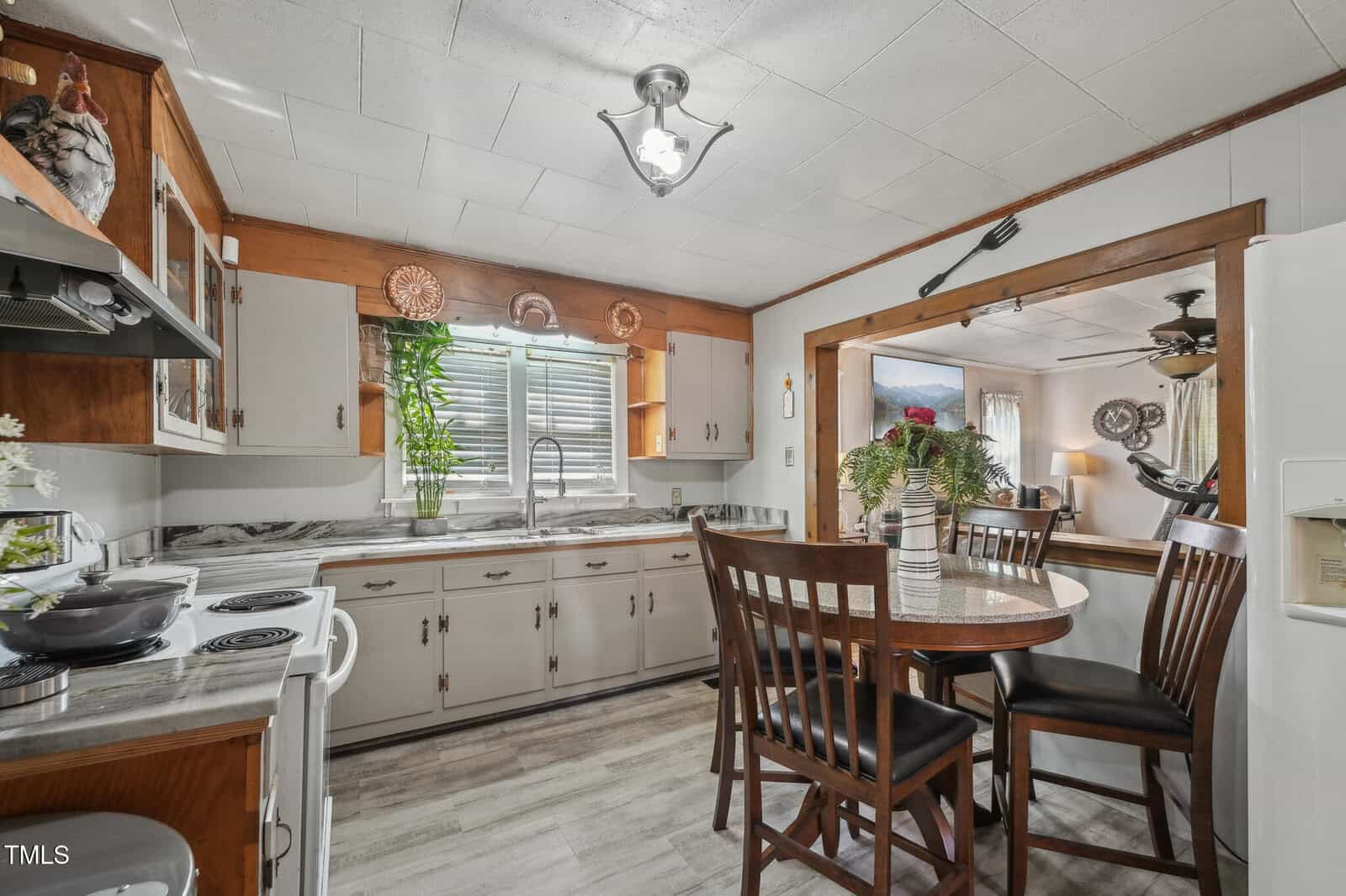 A kitchen with white cabinets, marble countertops, a round dining table with four chairs, plants by the window, and decorative plates on the wall. An open doorway connects to a living room area.