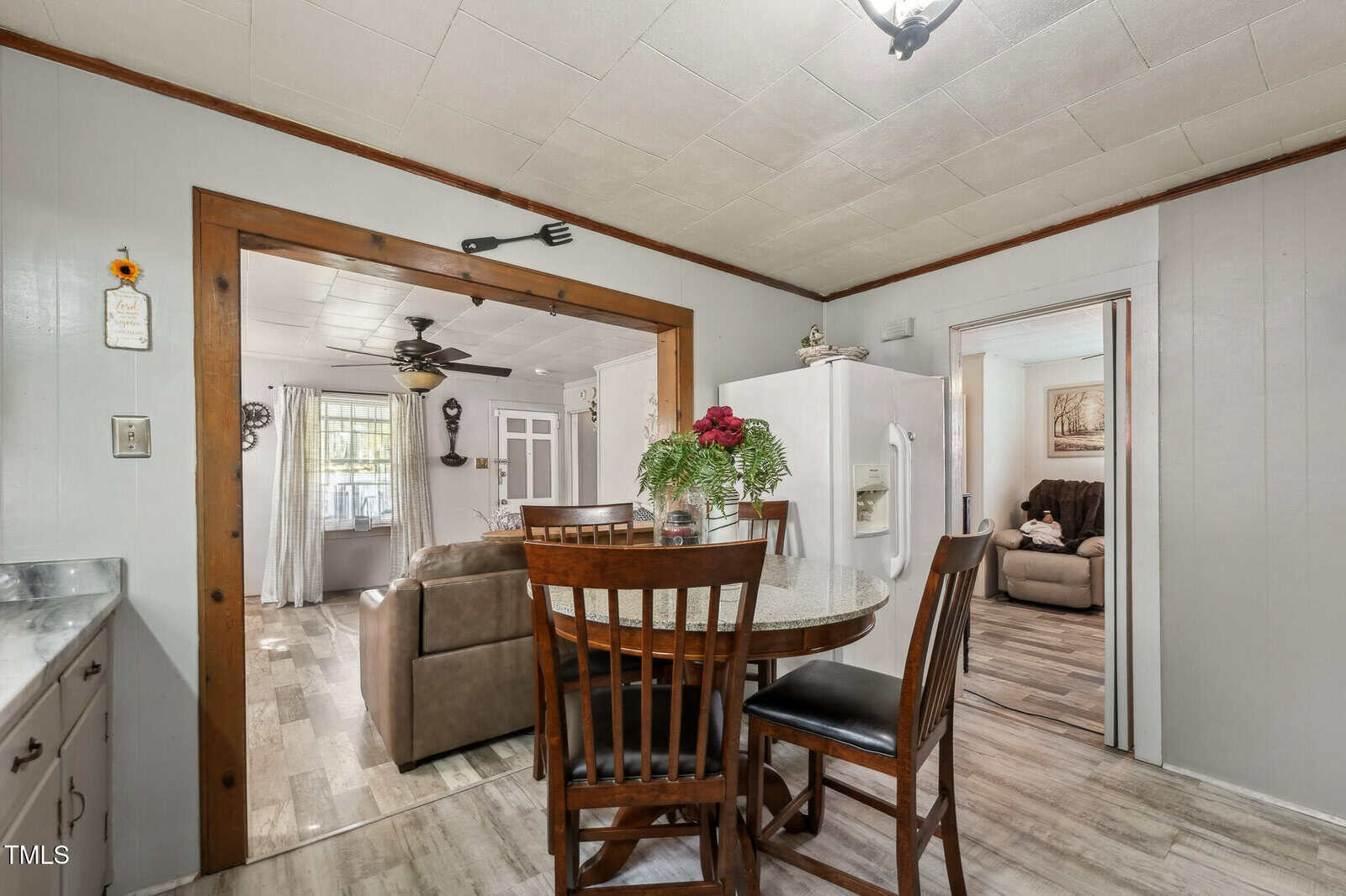 A kitchen area with a small round dining table and four chairs, a white refrigerator, and a view into a living room with a couch and ceiling fan, plus another room with a recliner. Wood trim frames the open doorway.