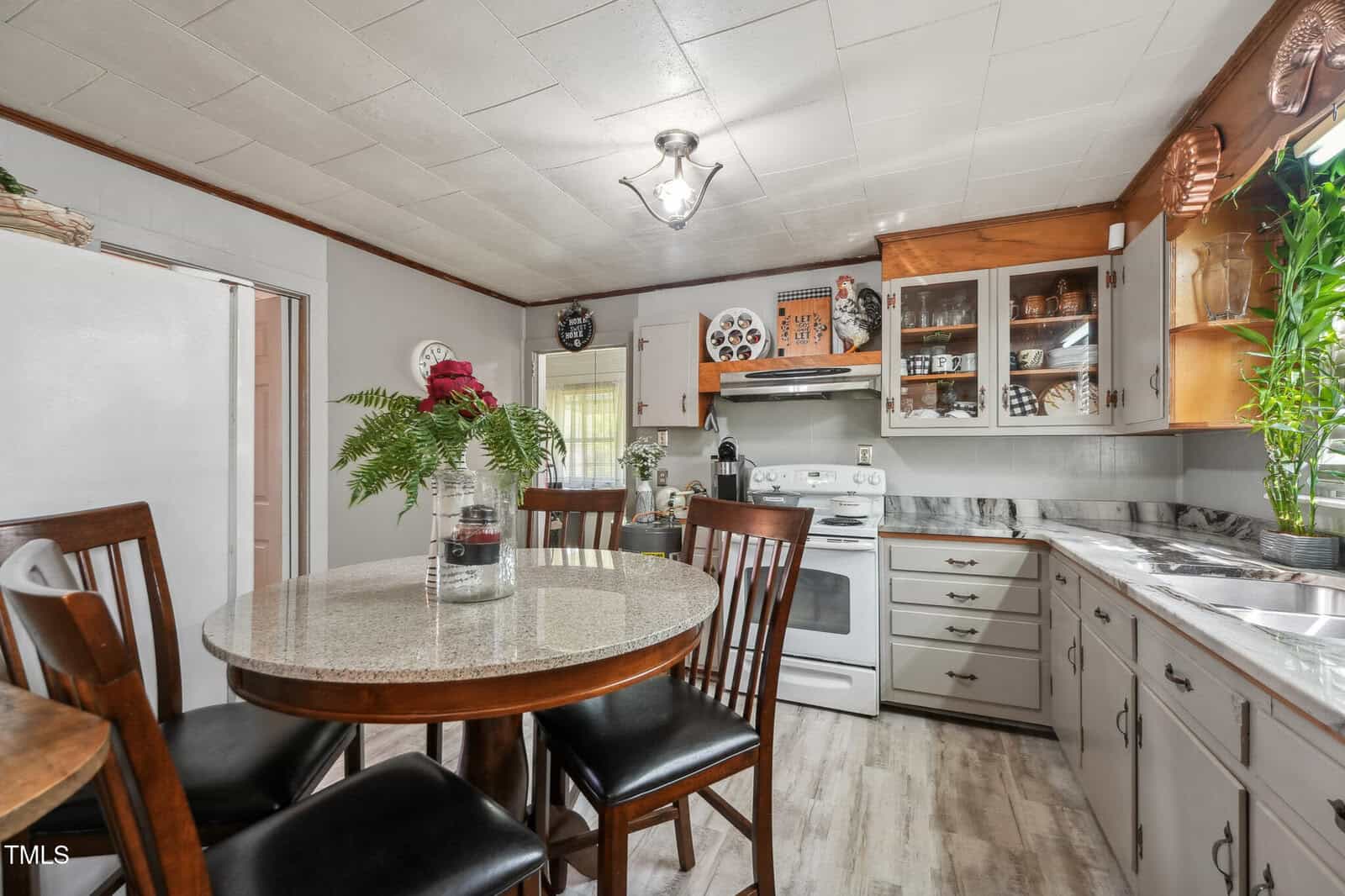 A kitchen with gray cabinets, a white stove and refrigerator, a round dining table with four chairs, and a plant centerpiece. Dishes and decor are displayed on shelves and the countertop near a window with green plants.