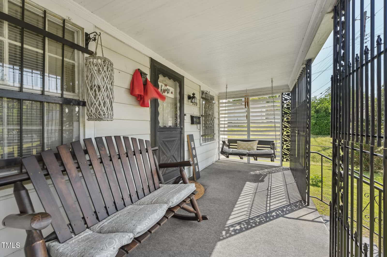 A covered front porch with two wooden rocking chairs, a swing, metal security doors and window bars, hanging lanterns, and a red cloth draped over a light fixture. Sunlight casts shadows across the carpeted floor.