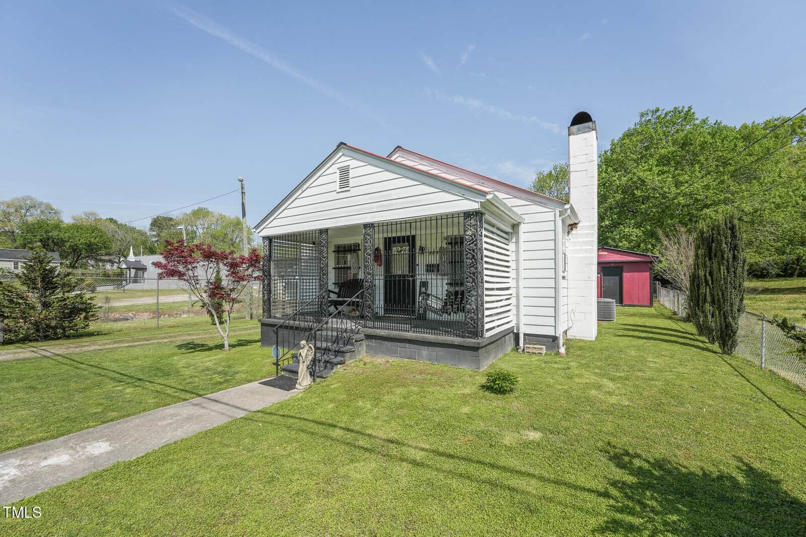 A small white house with a red roof, black railings, a covered front porch, and chimney stands on a grassy lawn with a concrete walkway. Trees and a red outbuilding are visible in the background.