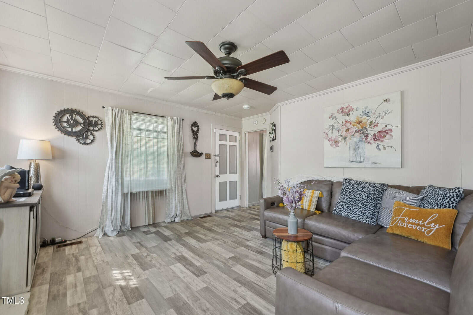 A living room with light wood flooring, white walls and ceiling, a gray sectional sofa with decorative pillows, a ceiling fan, a window with sheer curtains, and wall decor including a flower painting and clocks.