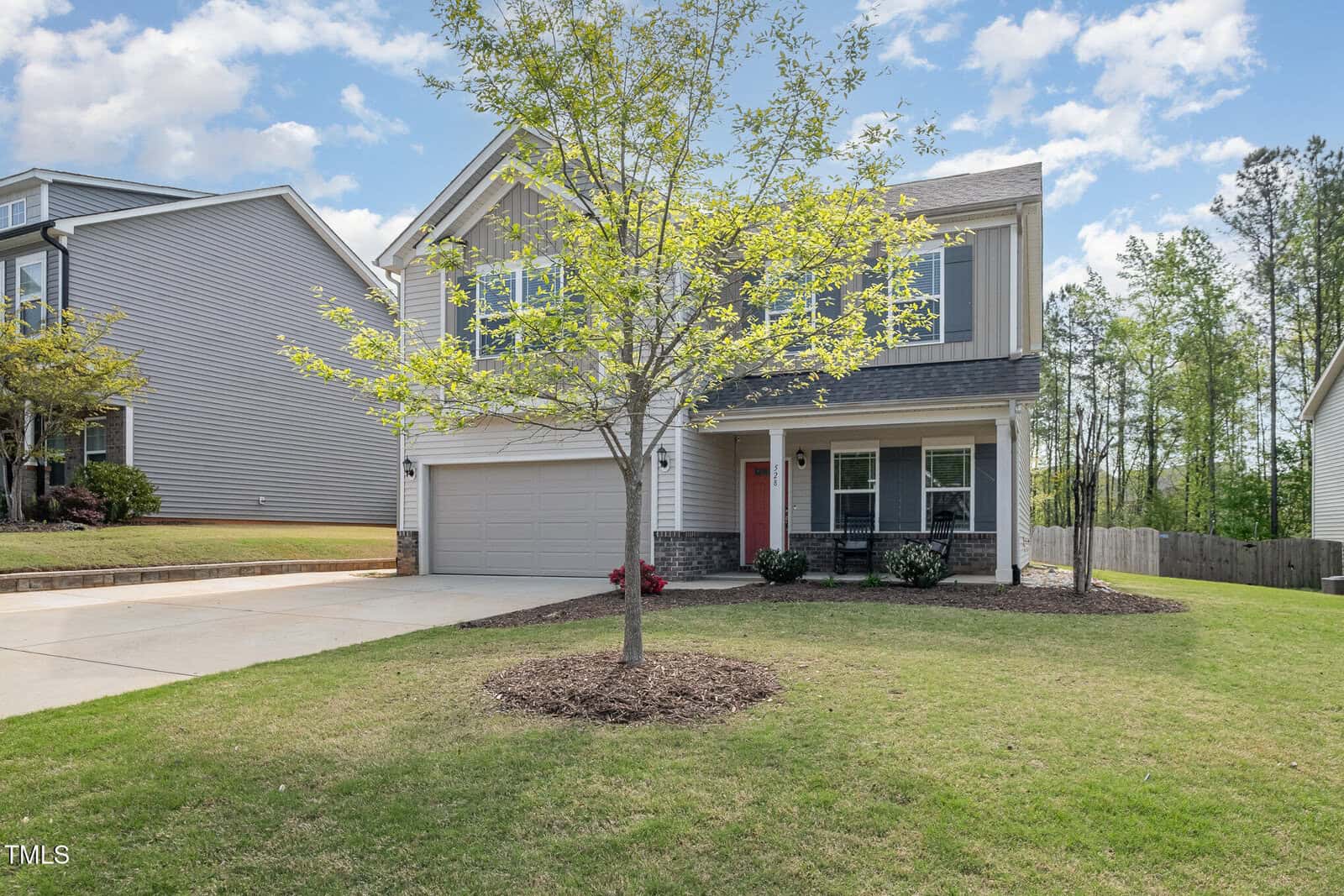 A two-story suburban house with a two-car garage, front porch, and light gray siding. A small tree and neatly trimmed lawn are in the front yard, with a wooden fence and trees in the background.