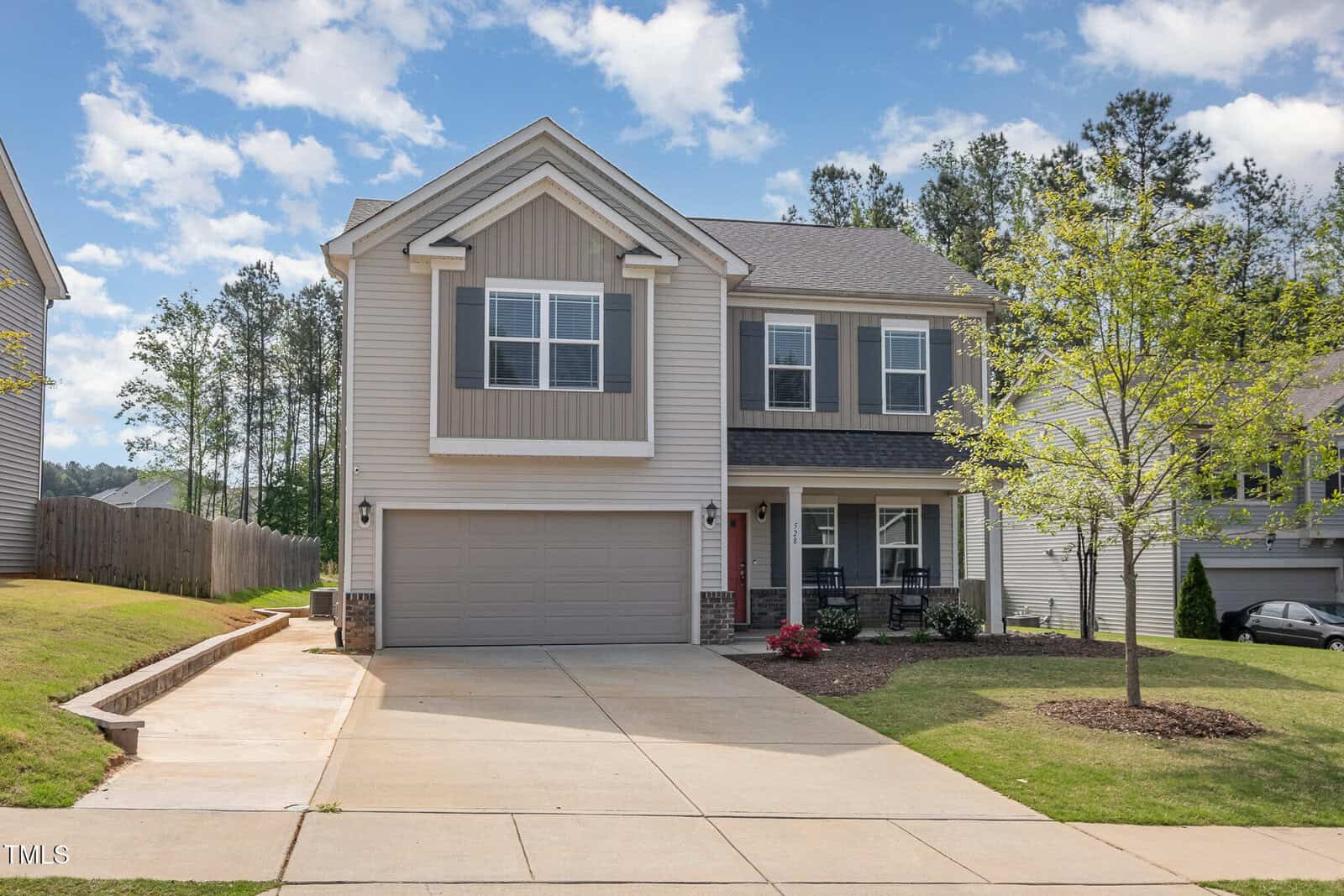 A two-story suburban house with gray siding, a double garage, and a paved driveway. There is a small front porch, neatly trimmed grass, and a young tree in the front yard. Trees line the background under a partly cloudy sky.