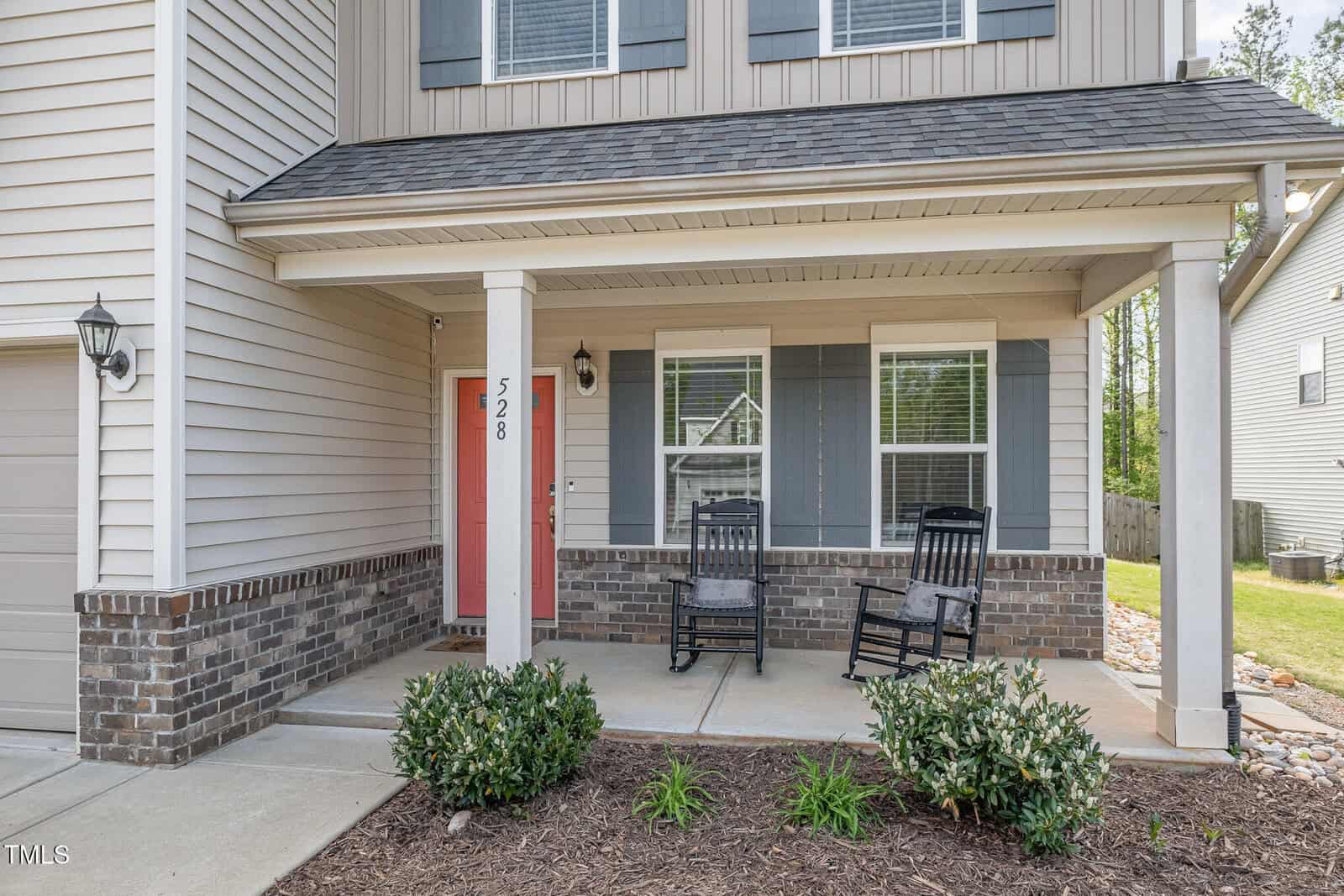 Front porch of a house with two black rocking chairs, a red front door labeled 528, brick and siding exterior, shrubs in front, and a window reflecting a neighboring home.