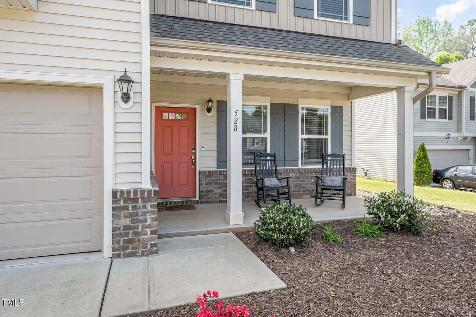 Front porch of a suburban house with a red door, brick and siding exterior, two black rocking chairs, some shrubs, and a concrete walkway leading to the entrance. A neighboring house is visible to the right.