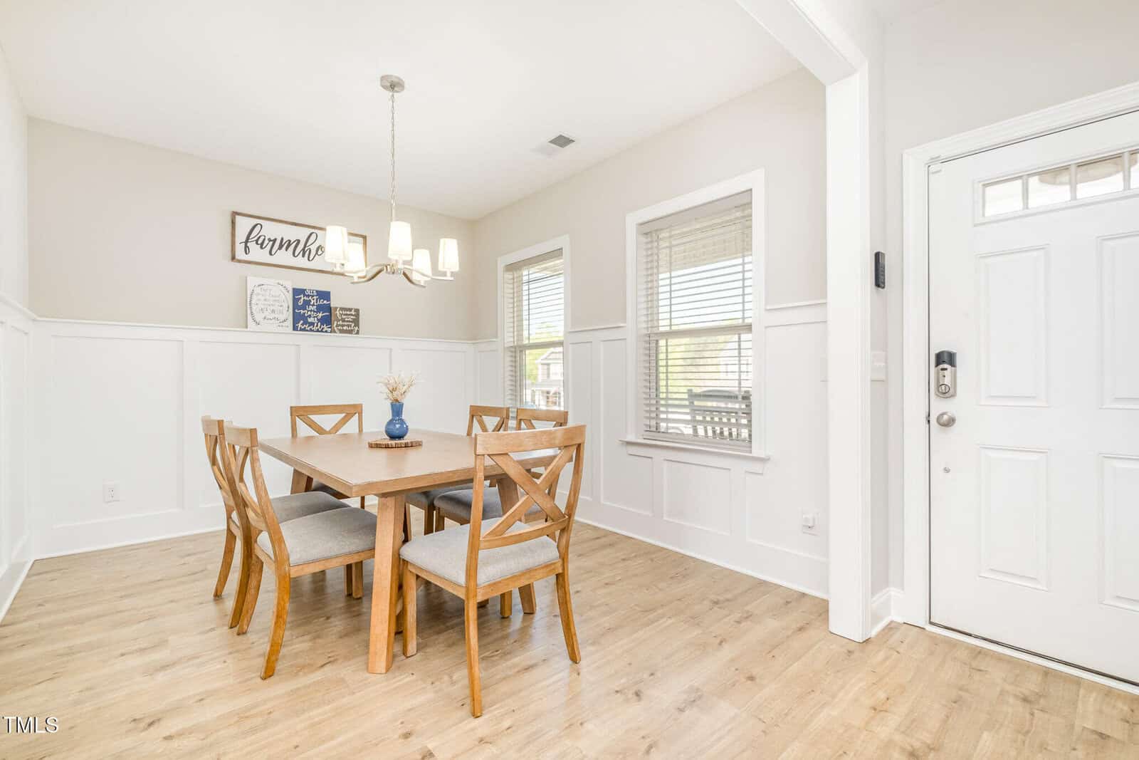 A dining area with a square wooden table and six matching chairs, light wood flooring, white walls with wainscoting, two large windows, a white door, and a modern chandelier overhead.