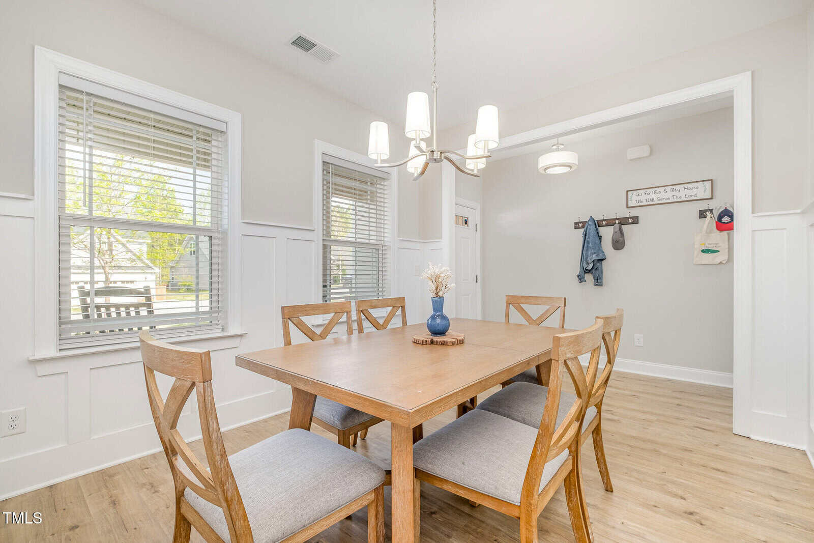 A dining area with a wooden table and six chairs is shown. Two windows with blinds, a modern chandelier, wood floors, and a small entryway with wall hooks are visible. Walls are white with vertical paneling.