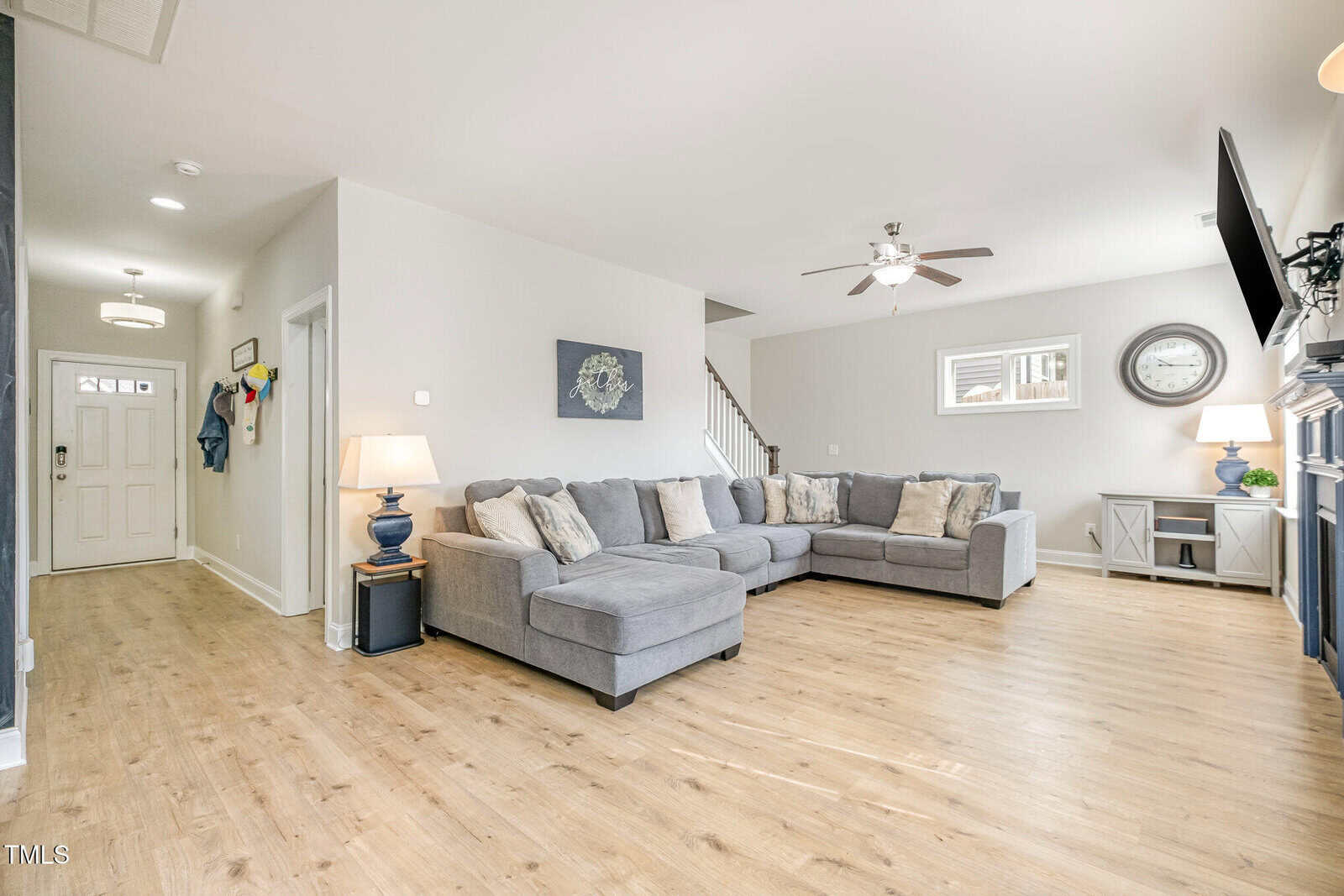 Modern living room with light wood floors, gray sectional sofa, blue lamps, wall art, and neutral walls. Front door and hallway are visible on the left, with a staircase and mounted TV on the right. Room is brightly lit.