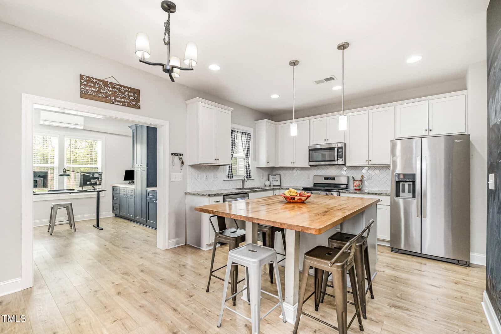 Modern kitchen with white cabinets, wood island with metal stools, stainless steel appliances, pendant lighting, and light wood flooring. An adjacent room with blue cabinetry and desk is visible through an open doorway.