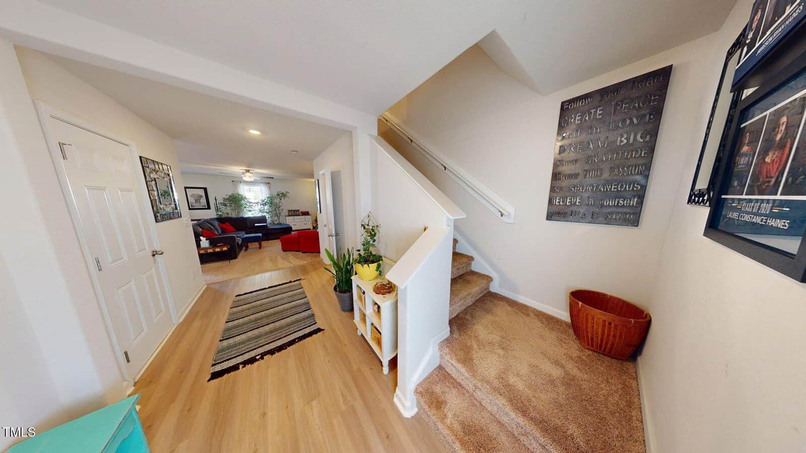 A hallway with light wood flooring leads to a carpeted staircase. To the left is a living room with couches. Plants, framed artwork, and a woven basket decorate the space. A rug lies near the entrance.