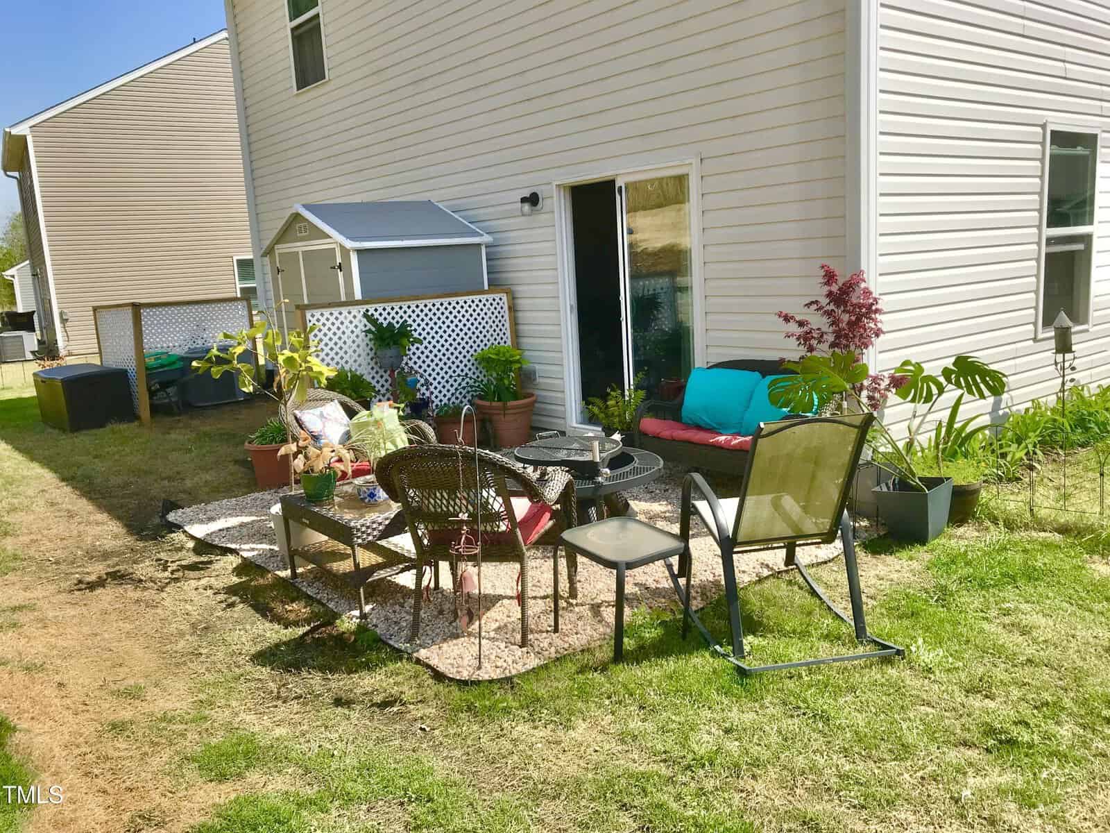 A backyard patio area with a rug, chairs, a small table, and potted plants arranged on a gravel section next to a beige house with sliding glass doors. There is a storage shed and neighboring homes visible.