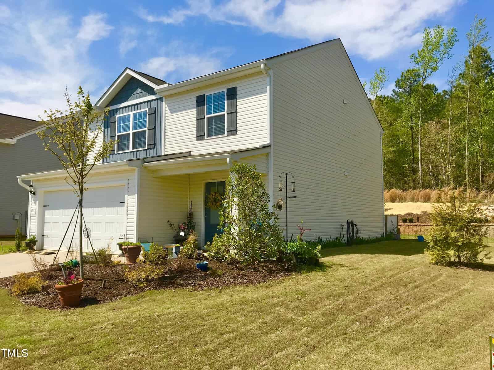 A two-story house with white siding and dark shutters, featuring a double garage, small covered porch, and landscaped front yard with young trees and shrubs. The lawn is neatly mowed, and trees line the background.