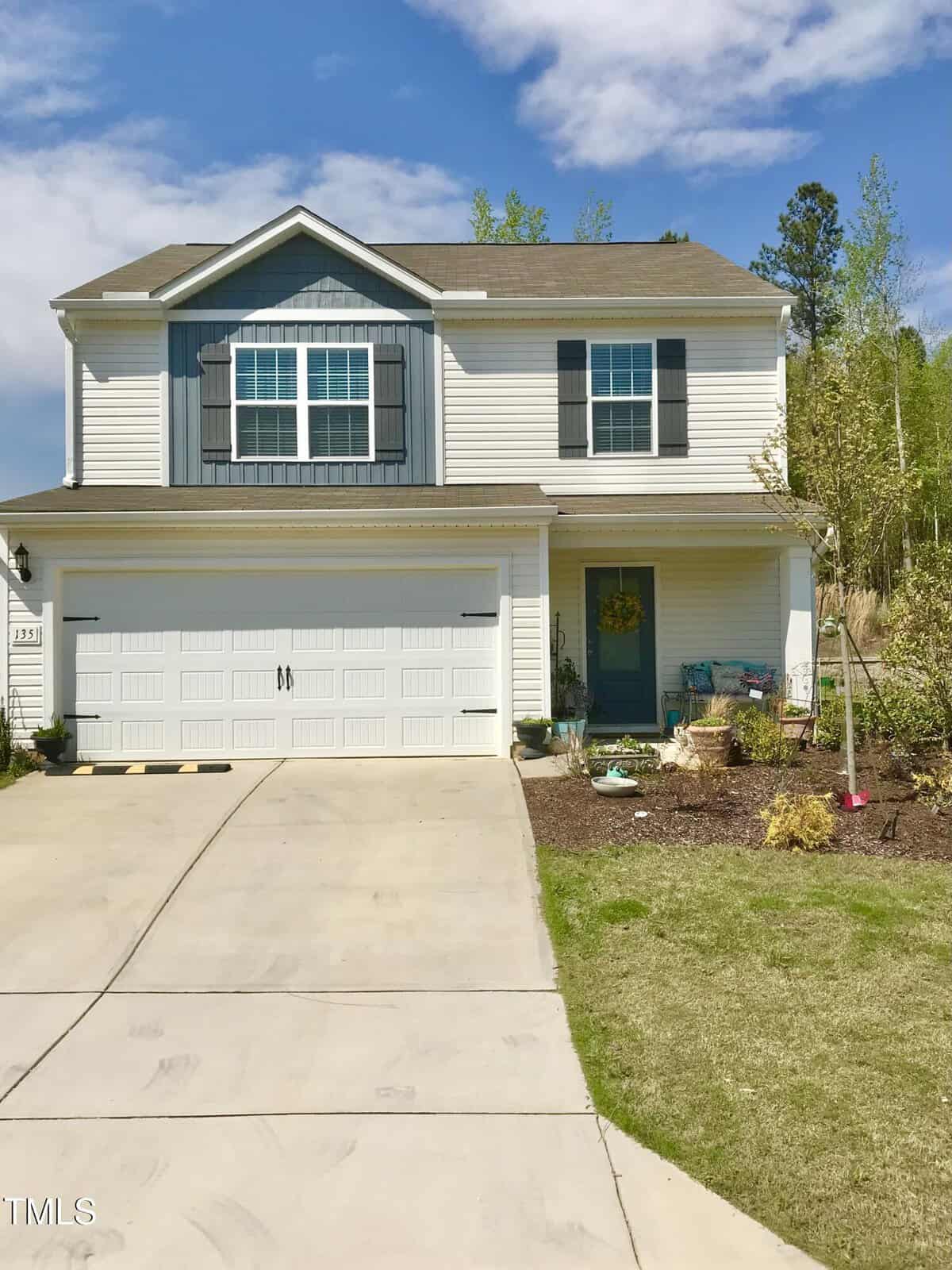 Two-story suburban house with white siding, gray shutters, and a double garage. A concrete driveway leads to the garage, and there are landscaped plants and a welcome wreath on the teal front door. Blue sky with clouds above.