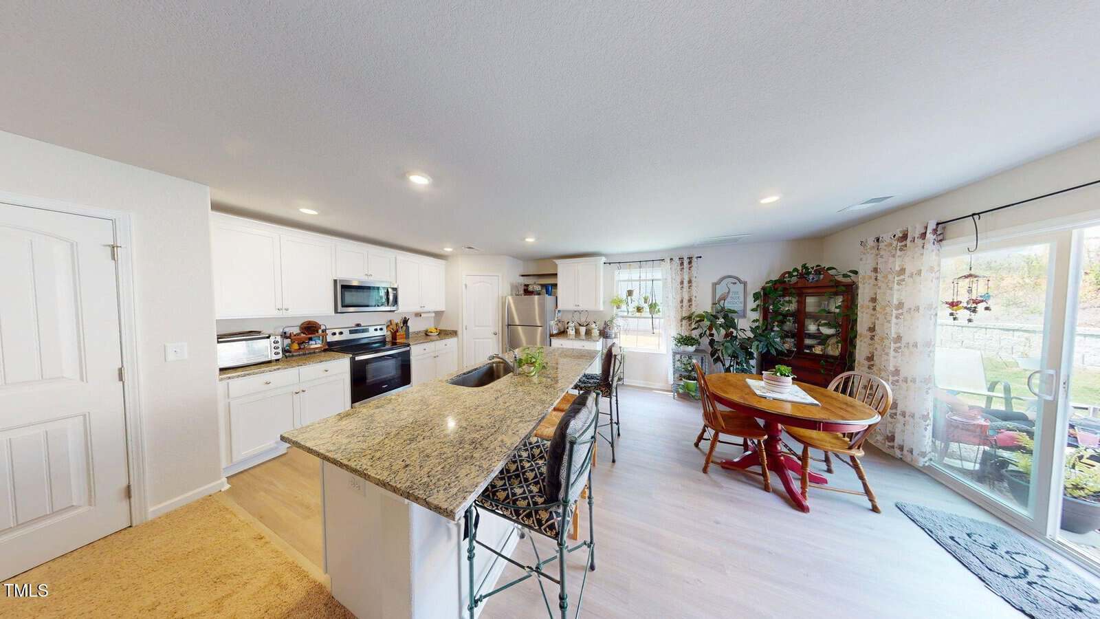 A modern kitchen with white cabinets, granite countertops, stainless steel appliances, an island with stools, and an adjacent dining area with a round wooden table, chairs, plants, and large windows with curtains.