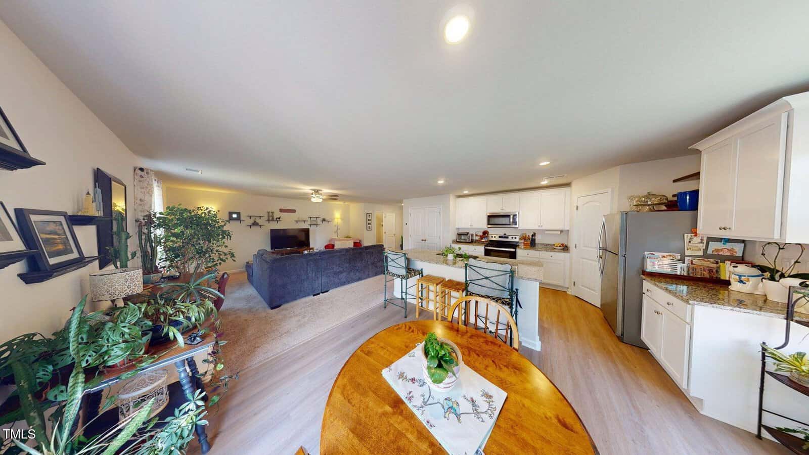 Open-plan kitchen and living area with white cabinets, stainless steel appliances, a round wooden dining table, green chairs, beige carpet in the living space, and houseplants on shelves and counters.