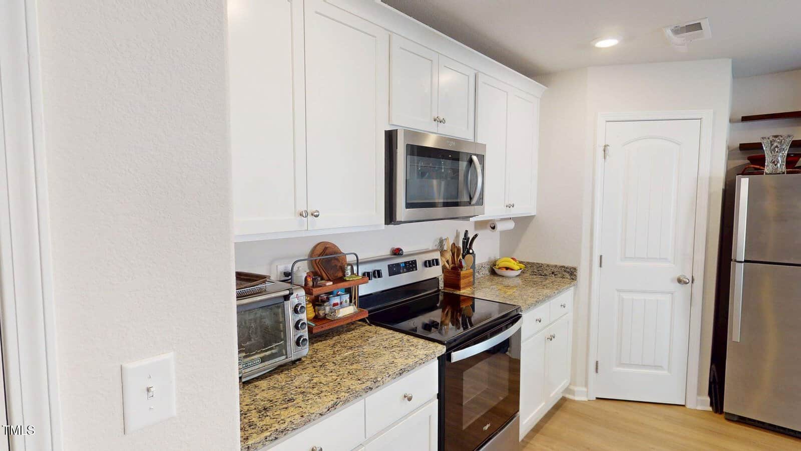 Modern kitchen with white cabinets, granite countertops, stainless steel appliances including an oven, microwave, and refrigerator, a toaster oven, utensils, and a fruit bowl. A white door and hardwood floor are visible.
