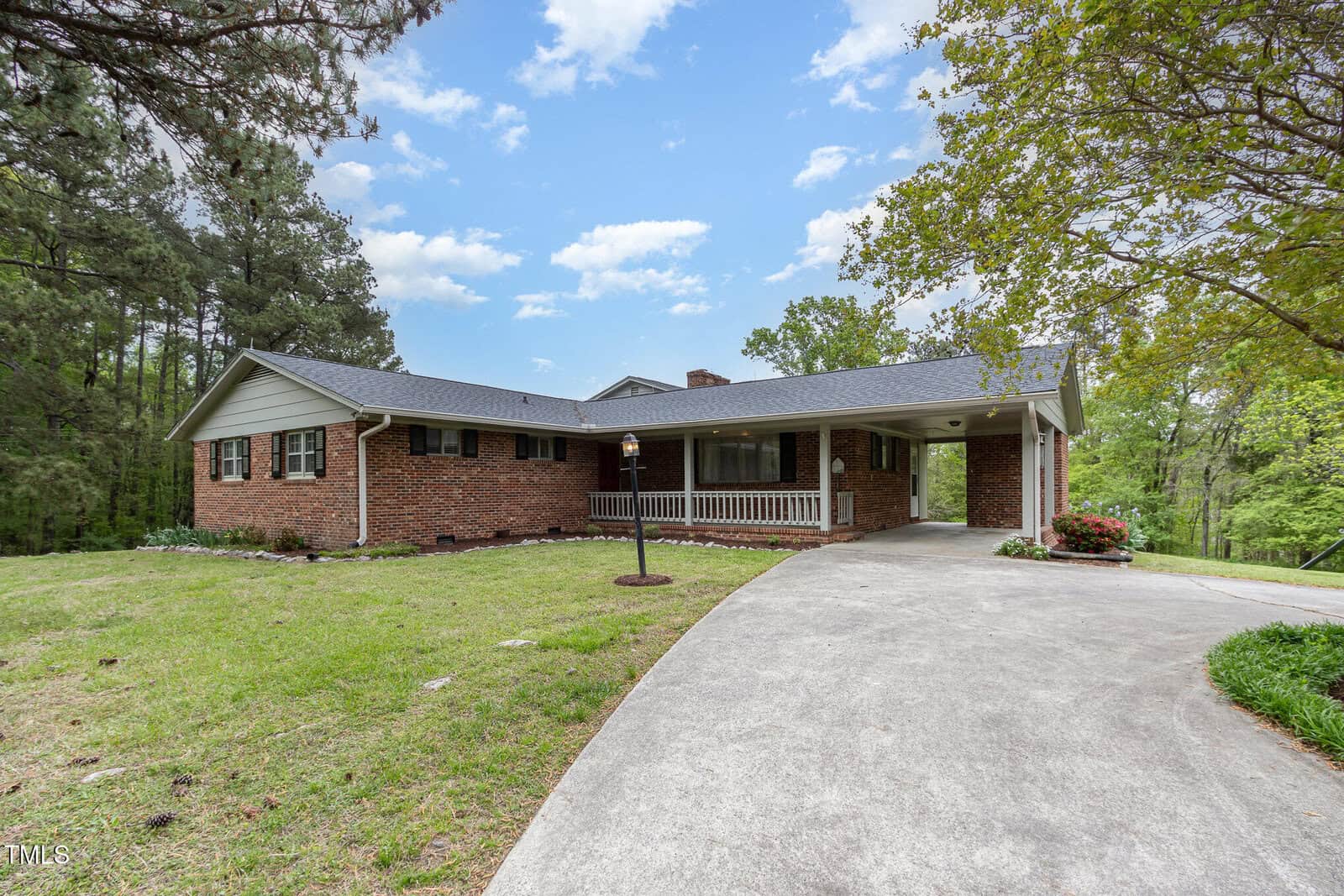 A single-story brick house with a covered front porch, white railings, and a sloped driveway. The house is surrounded by grass, trees, and a partly cloudy blue sky.