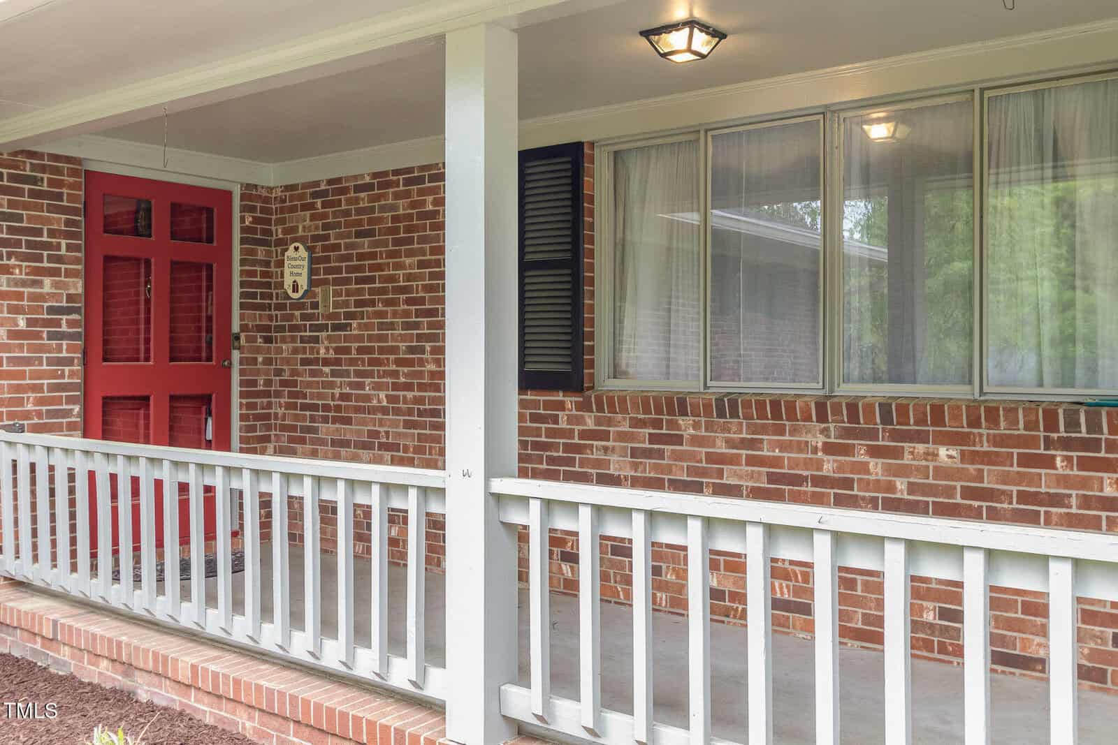A brick house front porch with a red door, white railing, and large windows with white curtains. The porch is covered with a ceiling light fixture and has a small step leading up from the walkway.