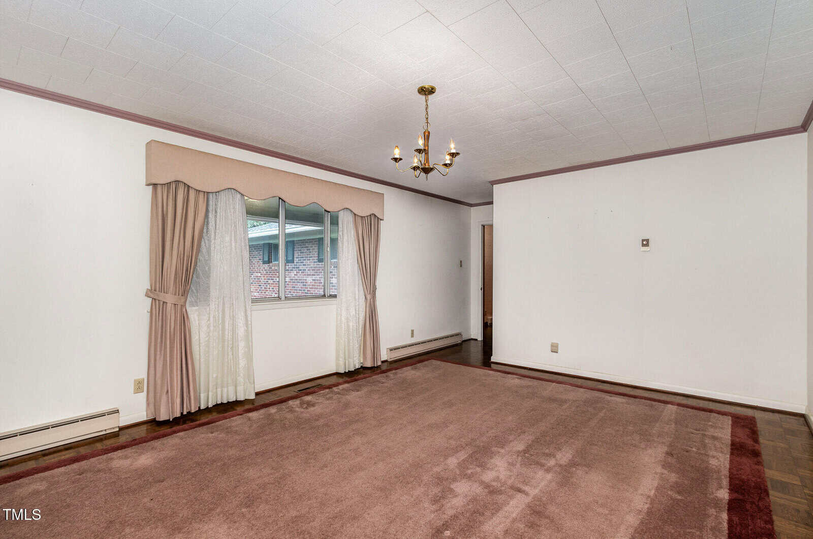 A mostly empty room with white walls, a light brown carpet, a window with beige curtains, a brass chandelier, and pink trim along the ceiling and baseboards. A doorway leads to another area.