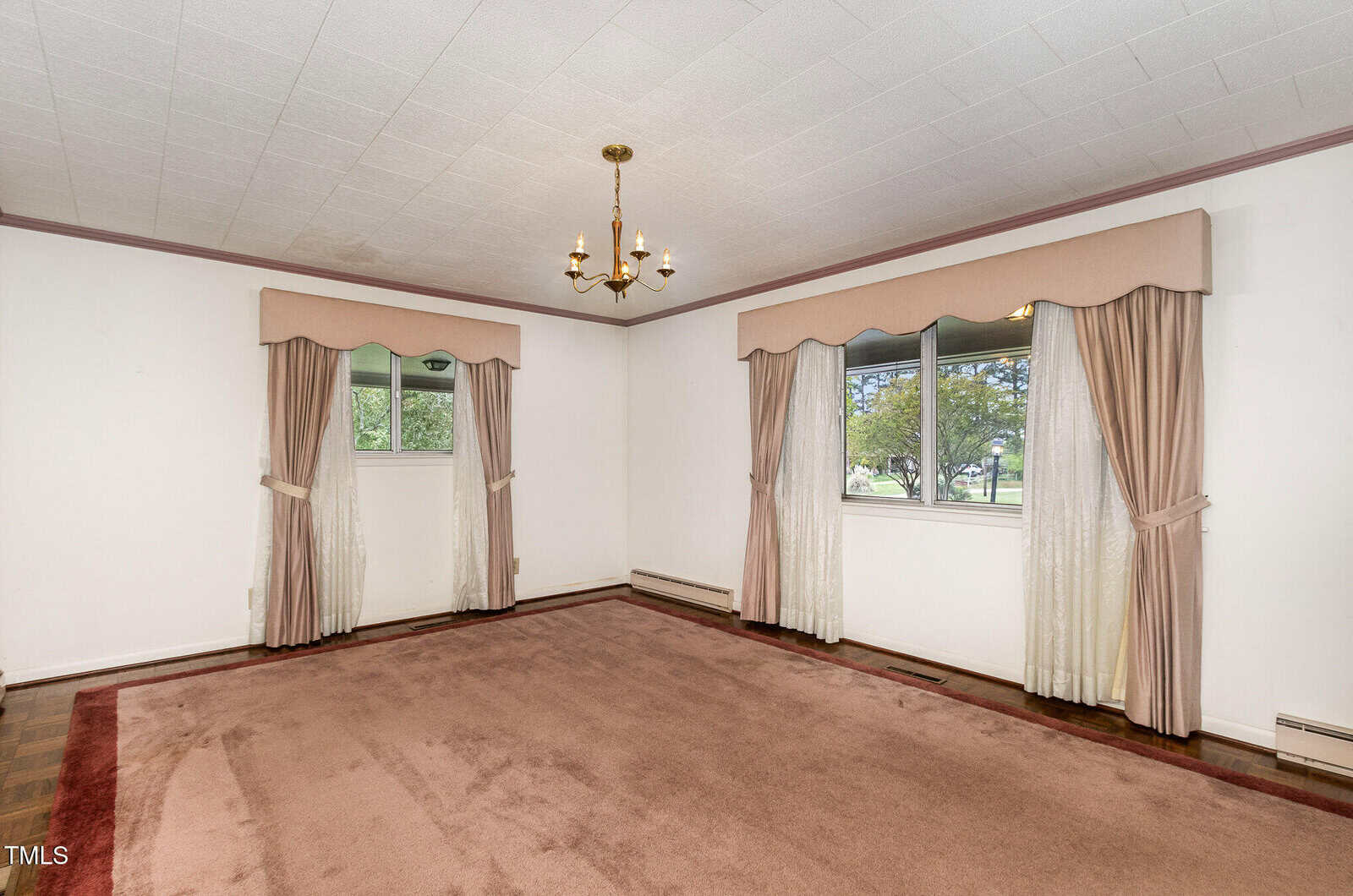 A mostly empty room with off-white walls, a beige carpet, two windows with drapes and valances, a brass chandelier, and baseboard heaters. The room has a ceiling with a grid pattern.