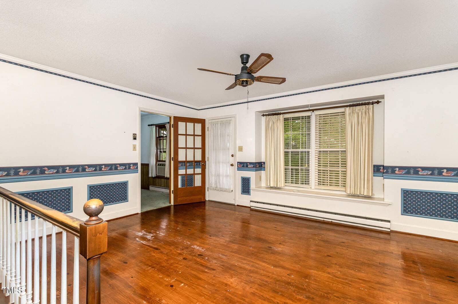 A spacious room with hardwood floors, white walls featuring blue duck-patterned wallpaper accents, a ceiling fan, large window with cream curtains, and a wooden door with glass panels leading to another room.