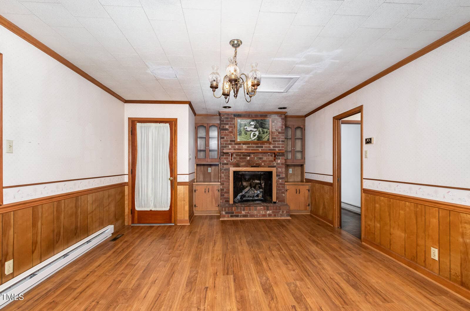 A room with wood panel wainscoting, a hardwood floor, a brick fireplace with built-in cabinets, a ceiling light fixture, and a wooden door with a glass panel. Ceiling has visible discoloration.