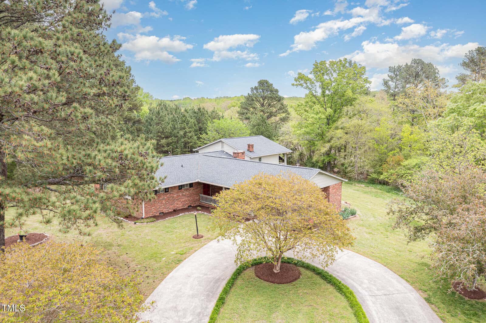 A large brick house with a gray roof sits among trees, surrounded by a circular driveway with mulched landscaping in the center. The sky is partly cloudy and the area is lush with greenery.