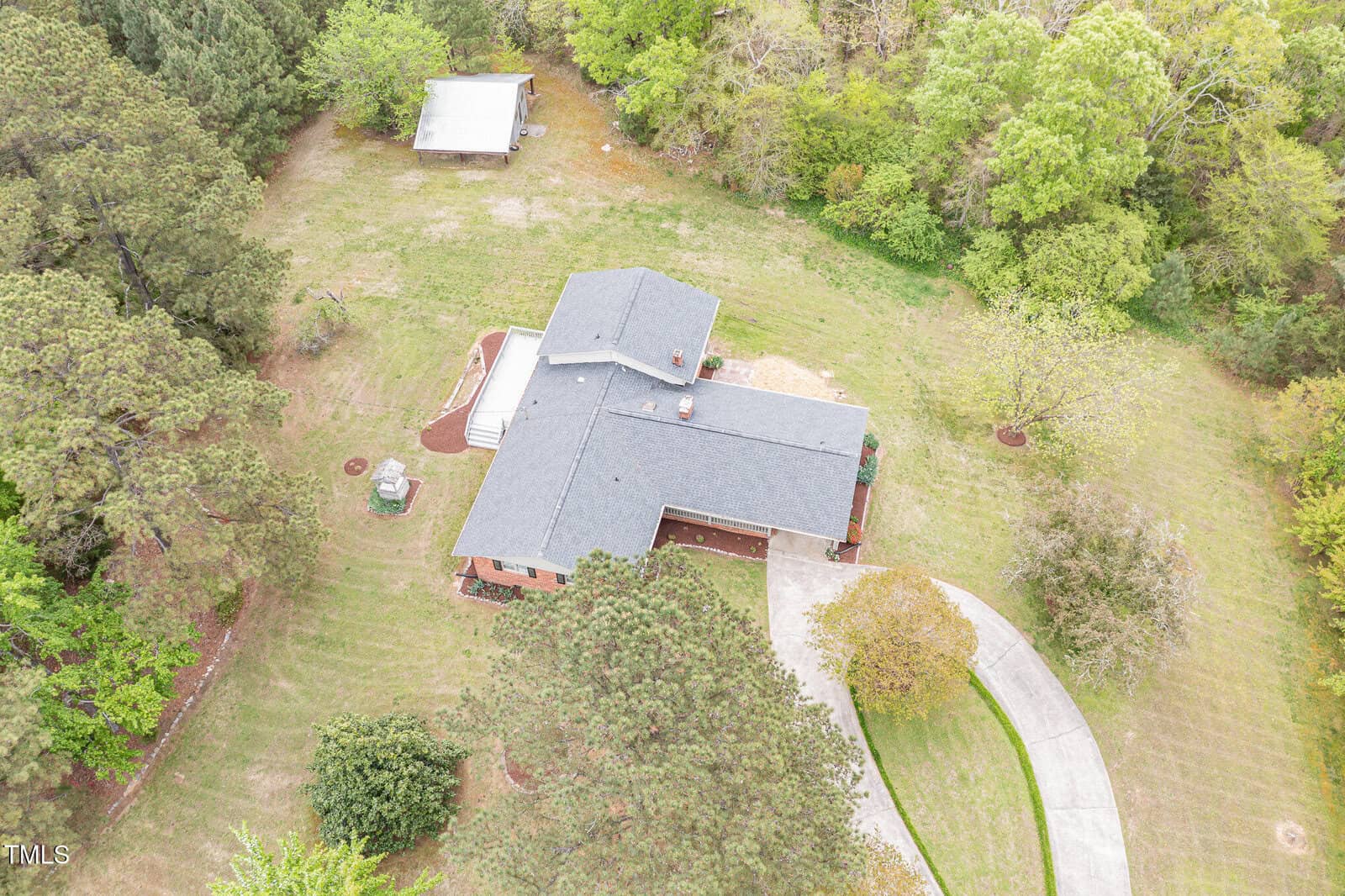 Aerial view of a single-story house with a gray roof, surrounded by trees and grass. A curved driveway leads to the front of the house, and a small detached building is visible in the background.