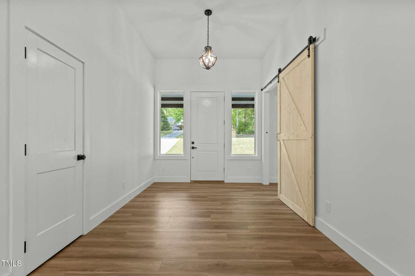 A bright entryway with white walls, wood flooring, a front door with glass panels, two side windows, a black pendant light, and a sliding wooden barn door on the right.